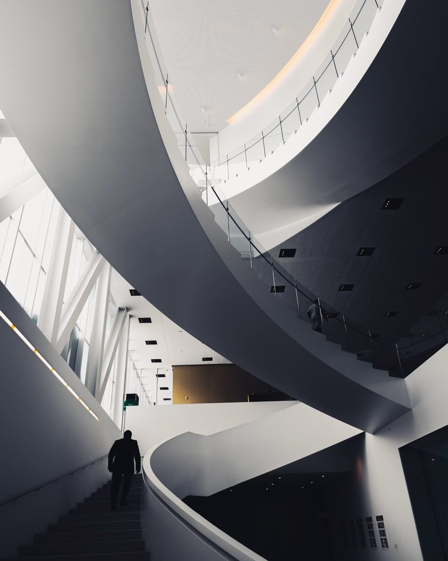 Escalier courbe dans un atrium blanc, une silhouette qui monte.