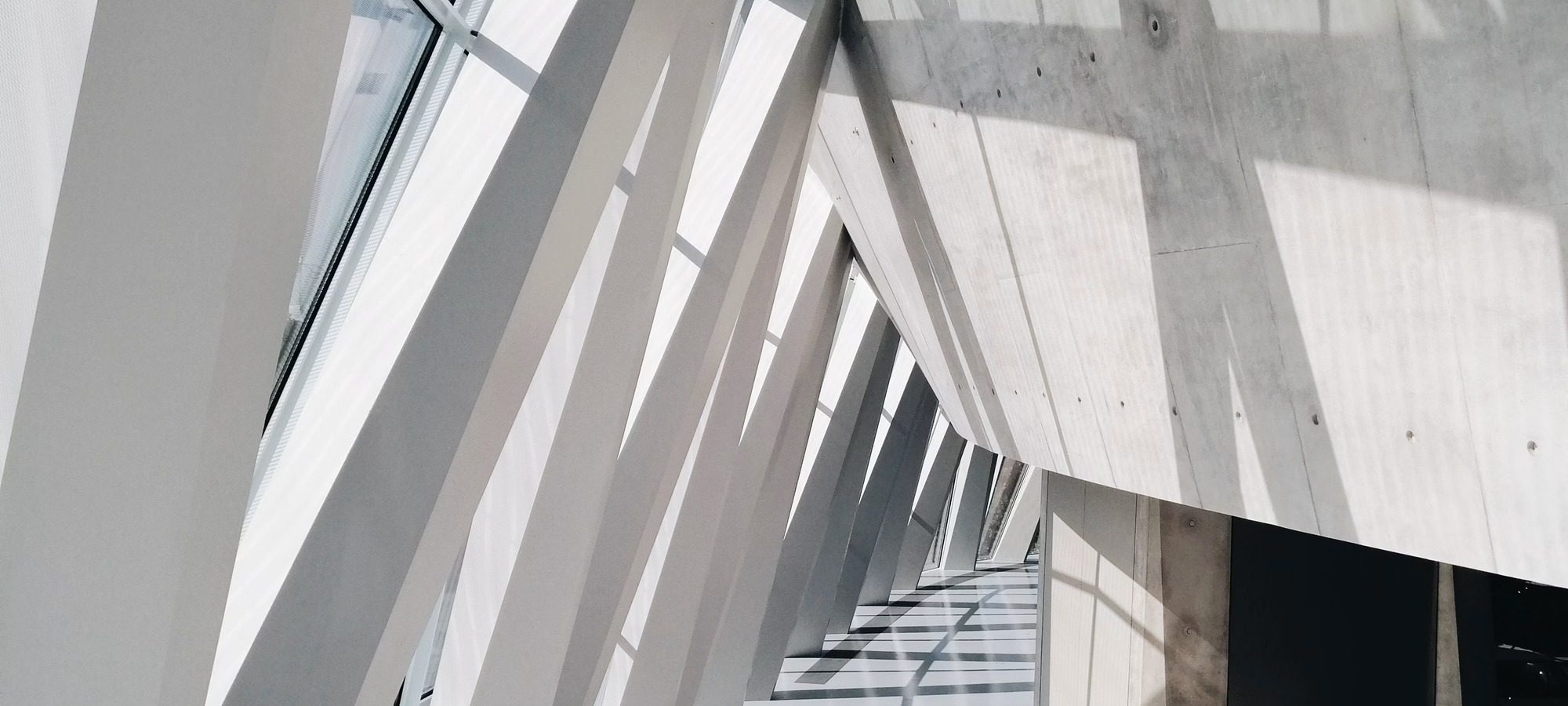 Atrium blanc, poutres obliques et escalier baignés de lumière.